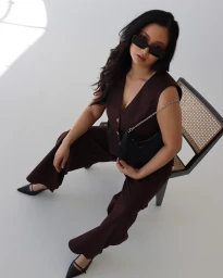 Woman in brown vest and trousers sitting on a cane chair wearing dark sunglasses and holding a black shoulder bag.
