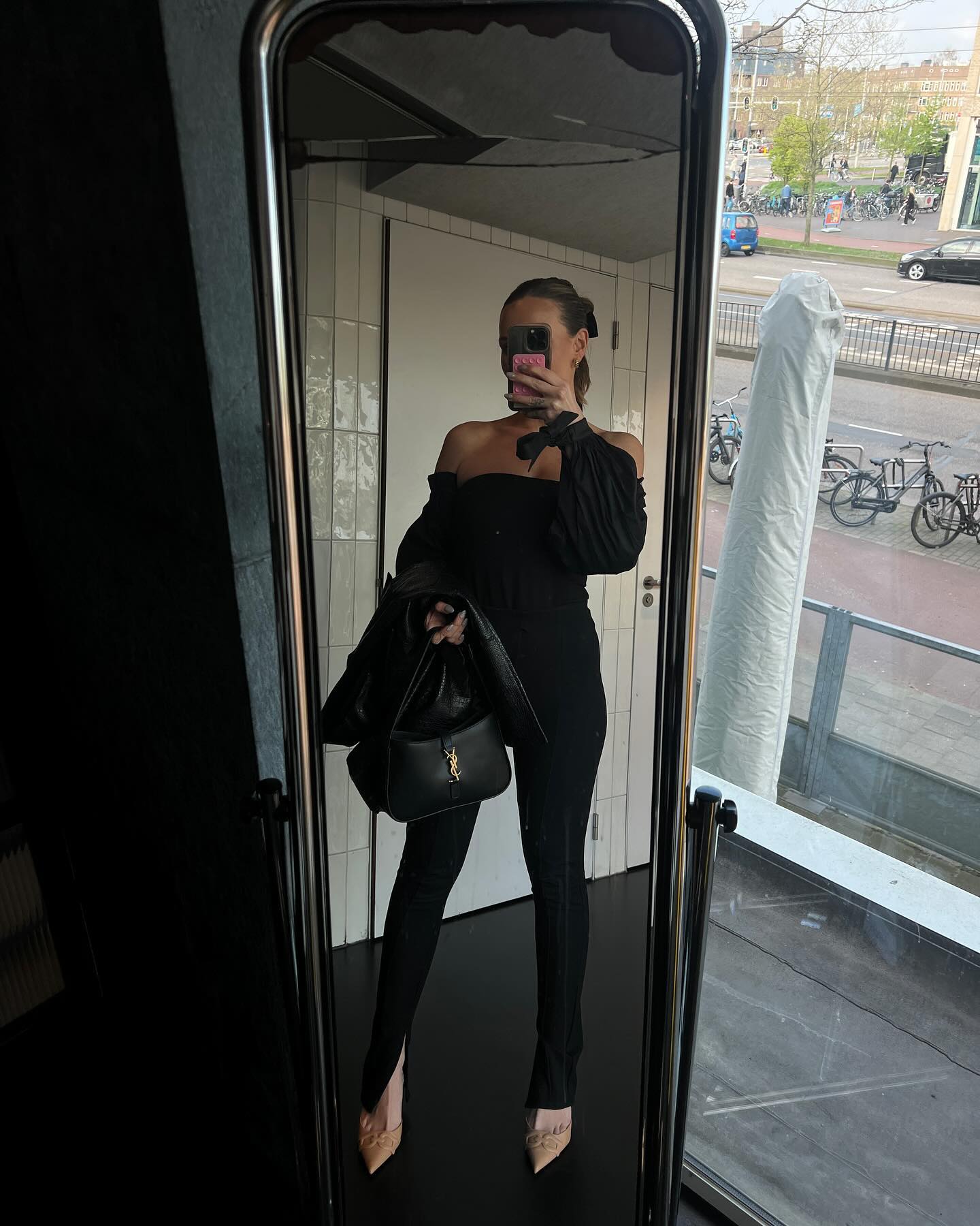 Mirror selfie of a woman in a black jumpsuit with slit pants and nude heels, holding a purse in an indoor setting with city view.