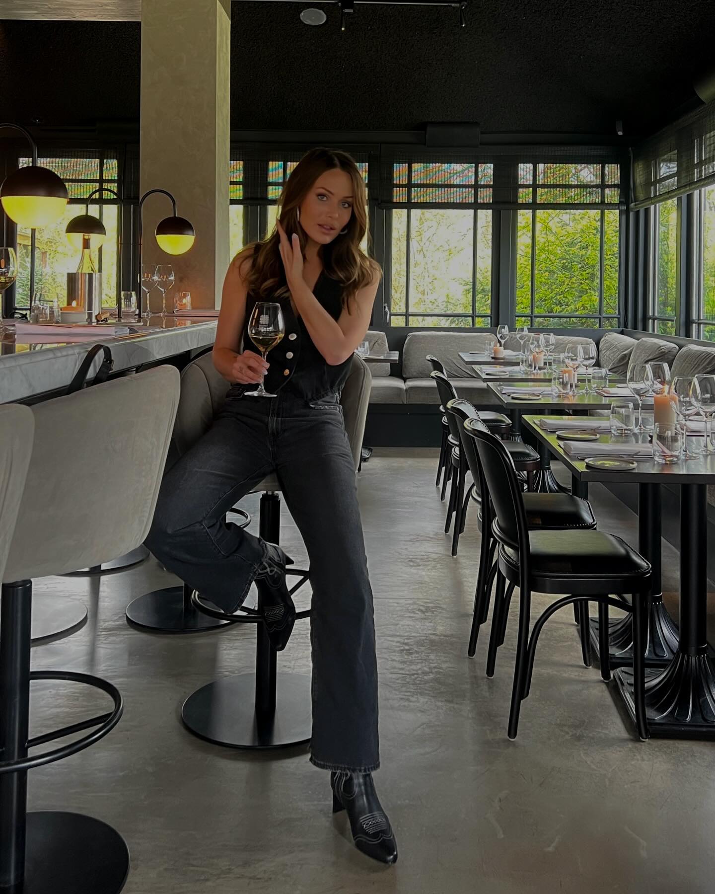 Adult woman in a sleeveless top and jeans sits on a bar stool holding a wine glass inside a restaurant with tables and chairs.
