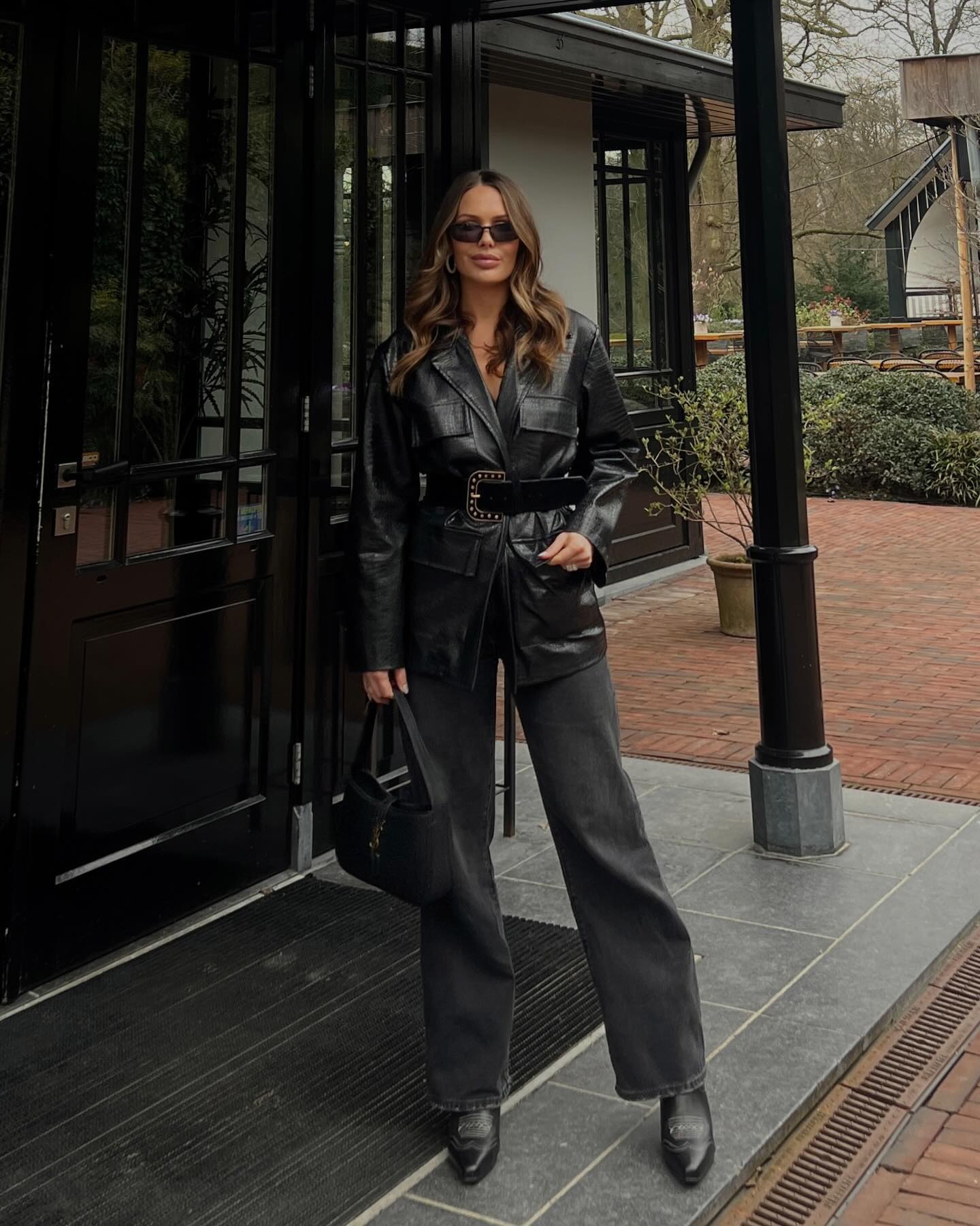 Brunette woman in a black leather jacket, wide-leg jeans, and heeled boots stands outside a building with black trim and red brick.