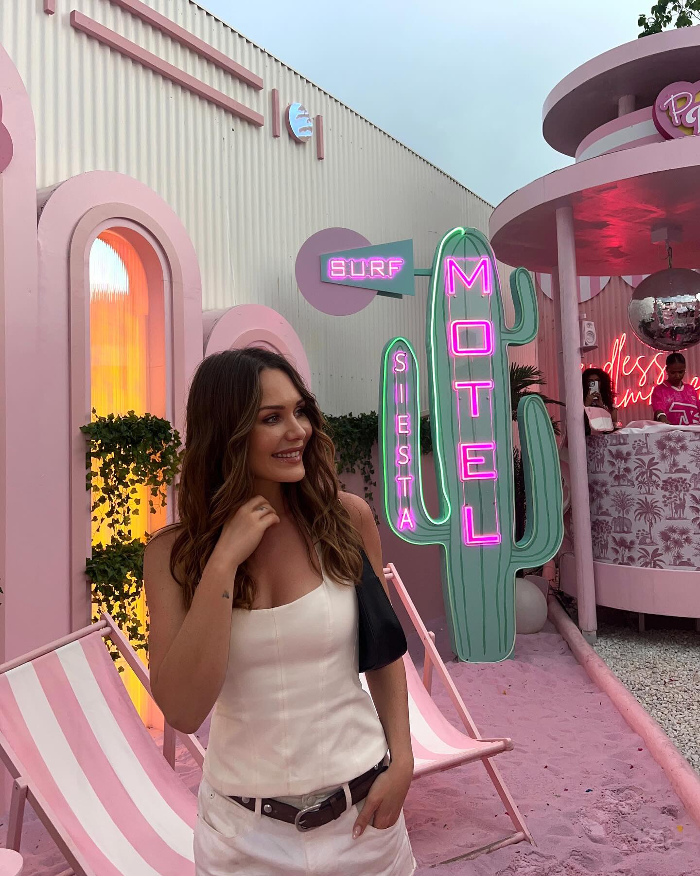 A young adult woman stands outdoors in front of a pink decorative building with surf and motel sign, with a chair in the foreground.