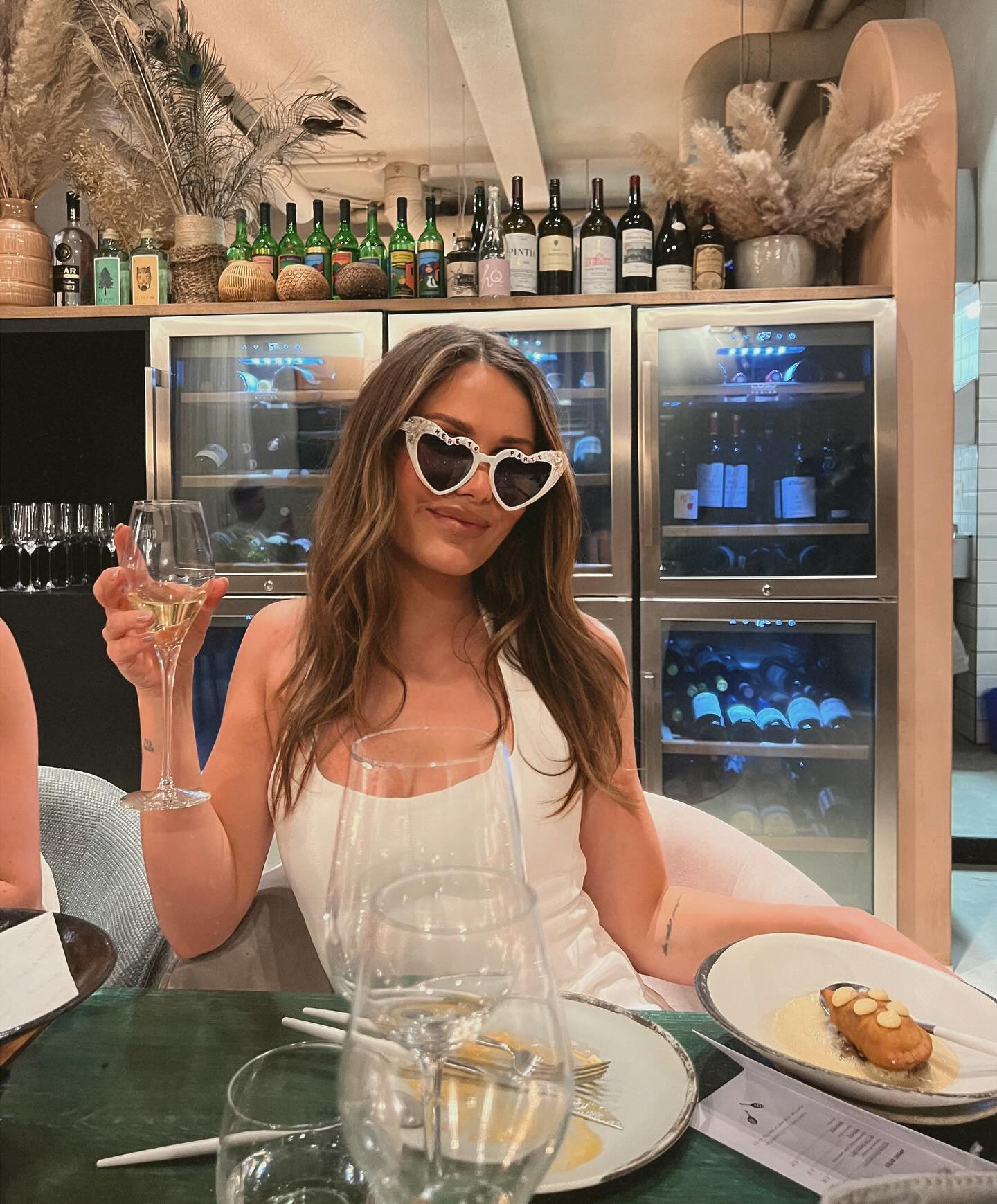Brunette woman in a white dress and heart-shaped sunglasses sits at a table with a glass of white wine inside a restaurant.