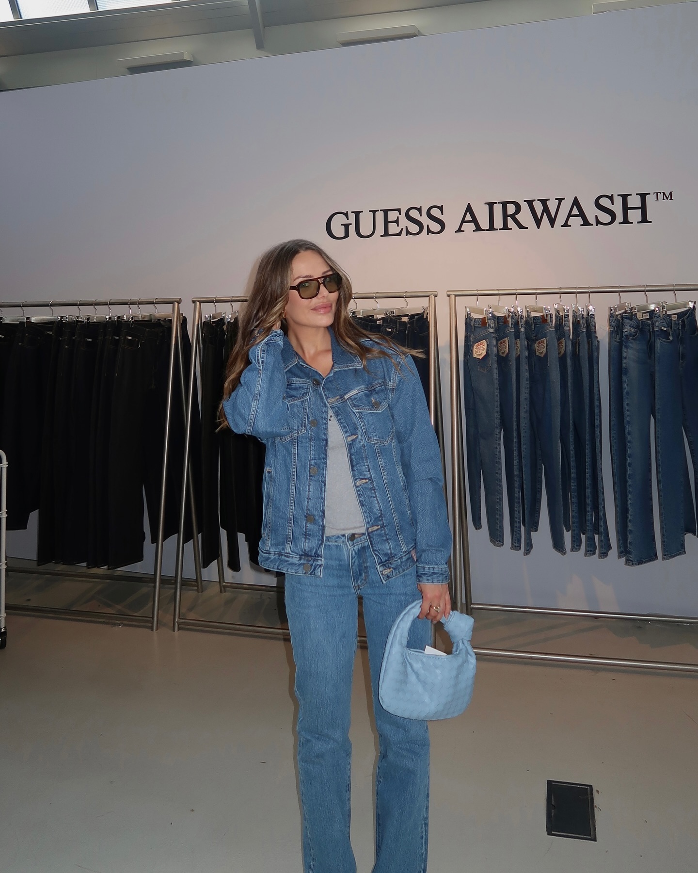 Woman in denim jacket and jeans with handbag stands in front of a rack of clothing at a clothing store, in a medium shot.