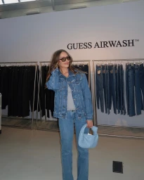 Woman in denim jacket and jeans holding a light blue handbag standing before clothing racks with Guess Airwash signage