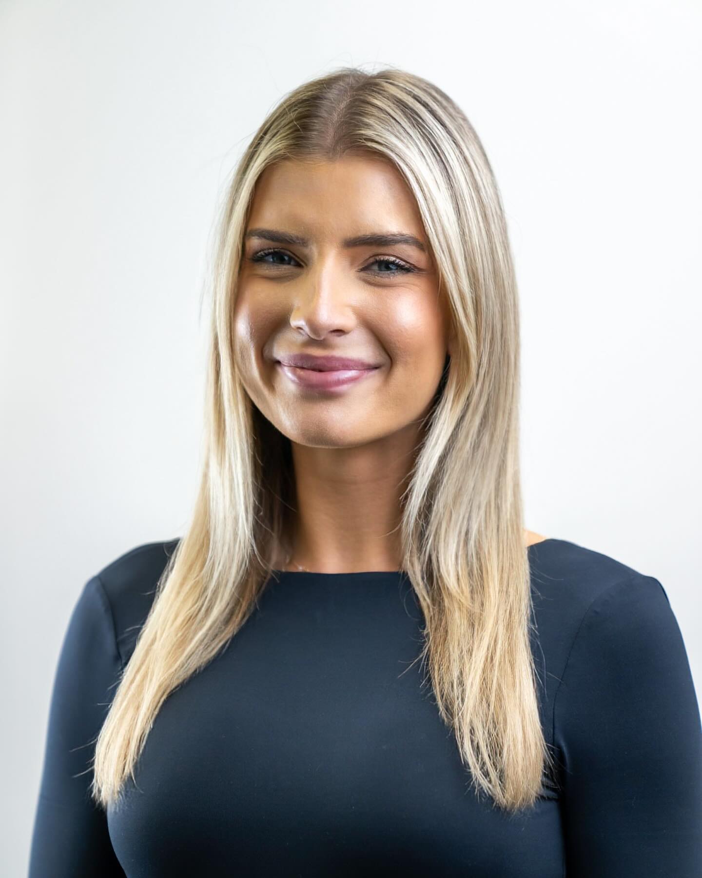 Portrait of a blond woman smiling, wearing a blue long-sleeve dress, shot in a bright indoor studio environment with neutral backdrop.
