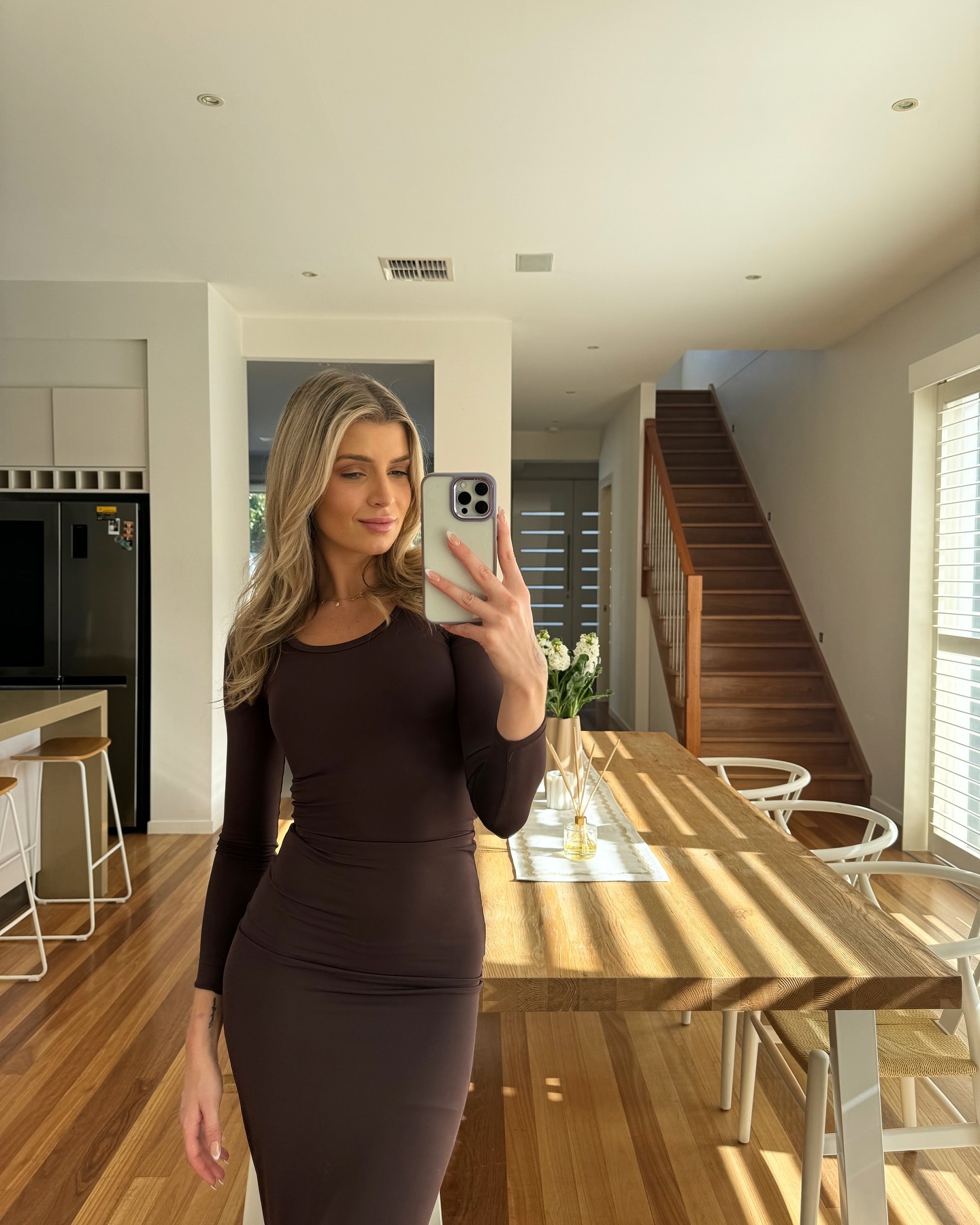 Blonde woman in brown dress takes a selfie in a home interior with a dining table, white flowers, and a staircase visible.