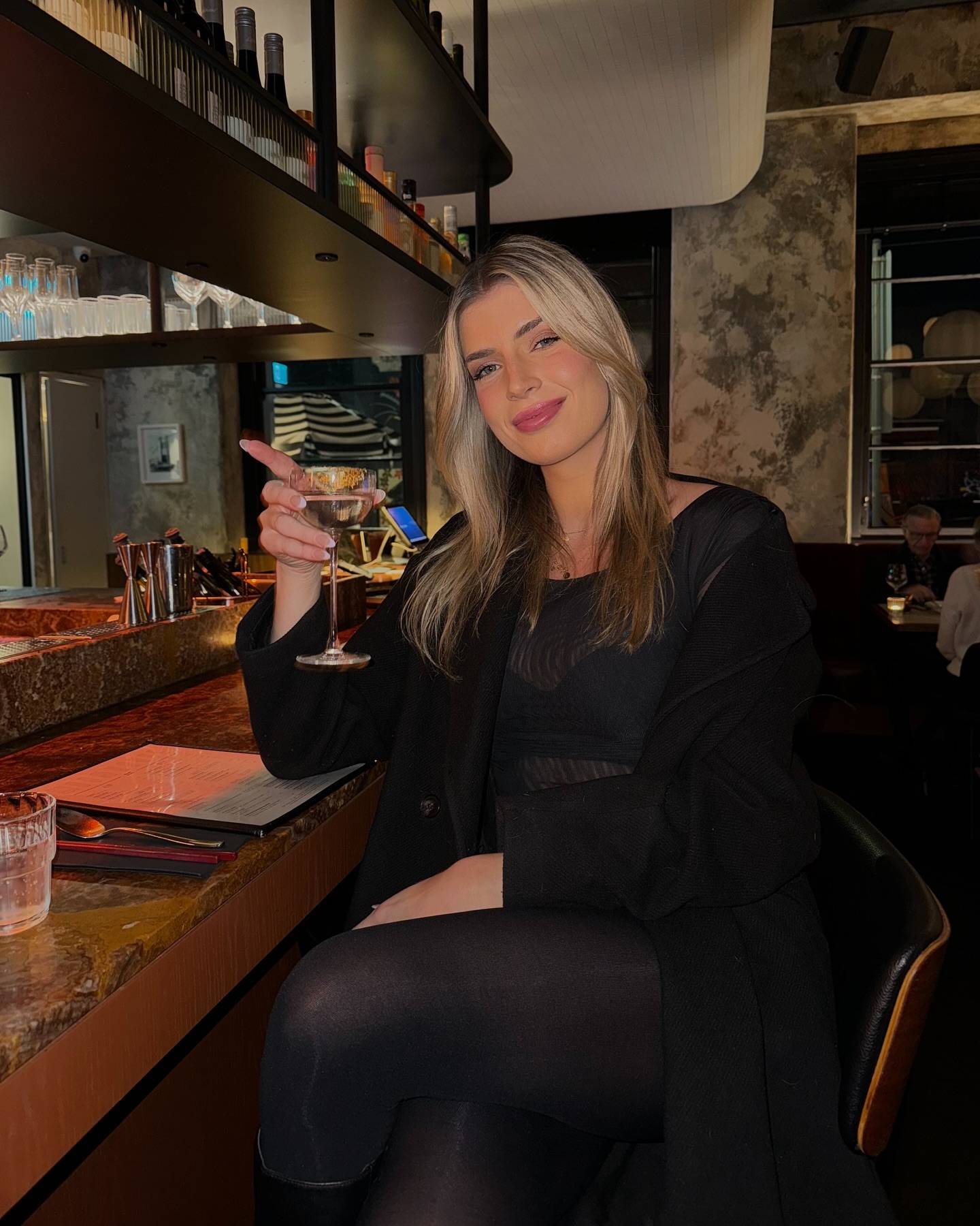 Woman in a black outfit poses indoors, holding a cocktail glass at a bar with drinks displayed on shelves in the background.