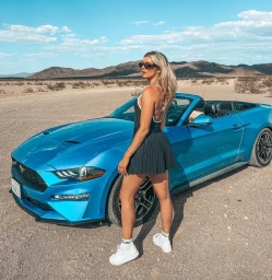 Woman in dark sunglasses and black miniskirt standing next to a bright blue convertible car in a desert landscape