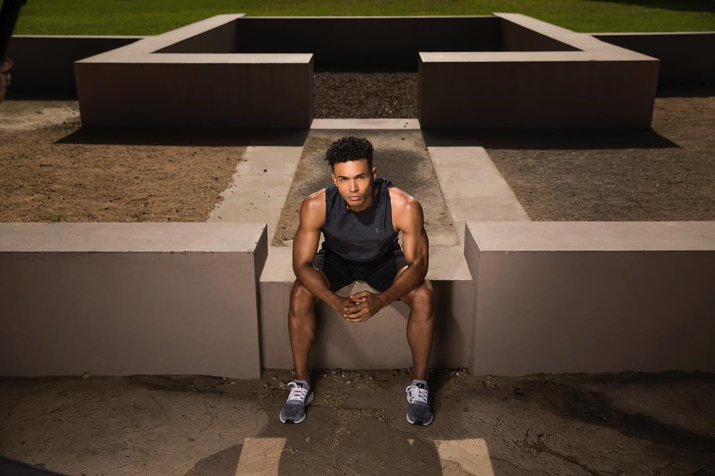 Athletic man in workout clothes sits on a concrete wall outdoors in neutral light. He wears a sleeveless shirt and athletic shoes.