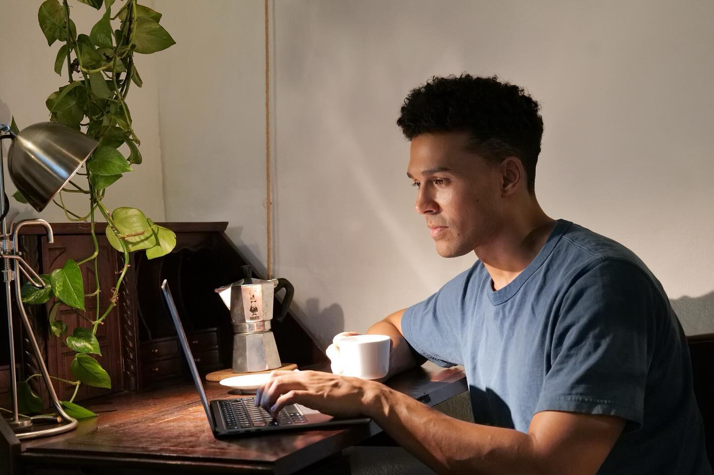 Man works on a laptop at wooden desk, with a coffee cup, moka pot, and desk lamp. Potted plant adds a natural element to workspace.