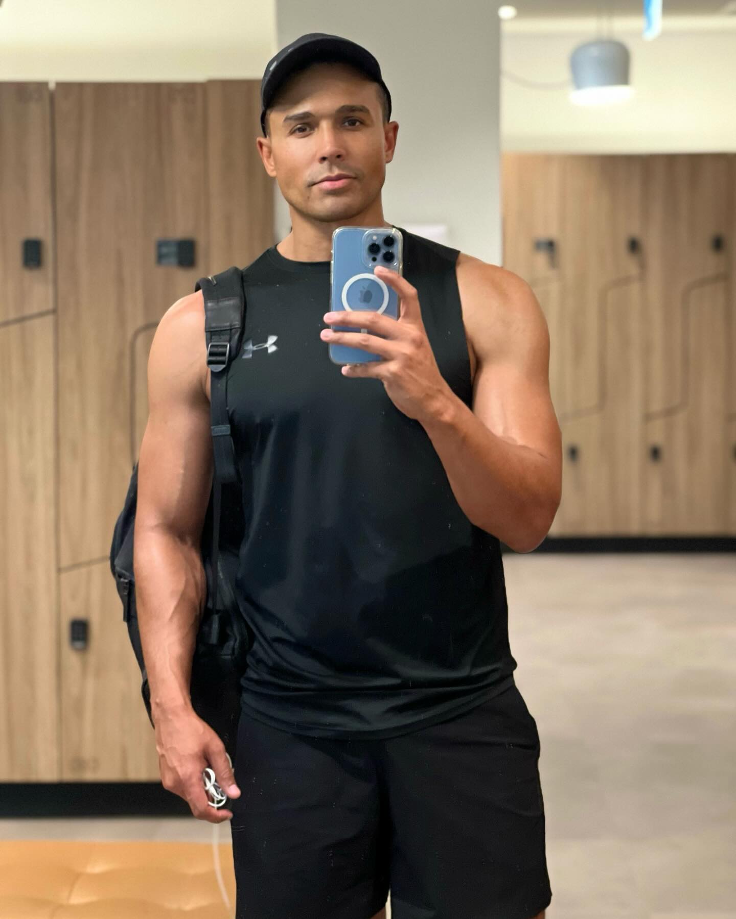 Athletic adult man in a sleeveless black shirt takes a selfie in a gym with lockers, holding a smartphone while wearing a baseball cap.