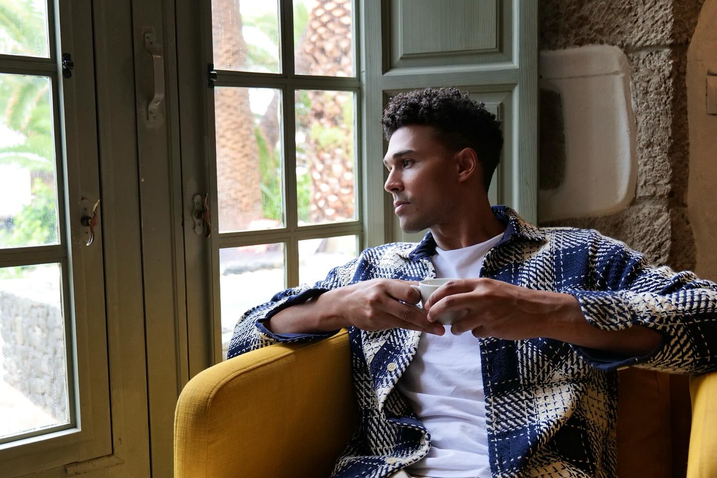 Indoor portrait of a man seated in a yellow chair, holding a cup, and looking out a window in a home interior setting.