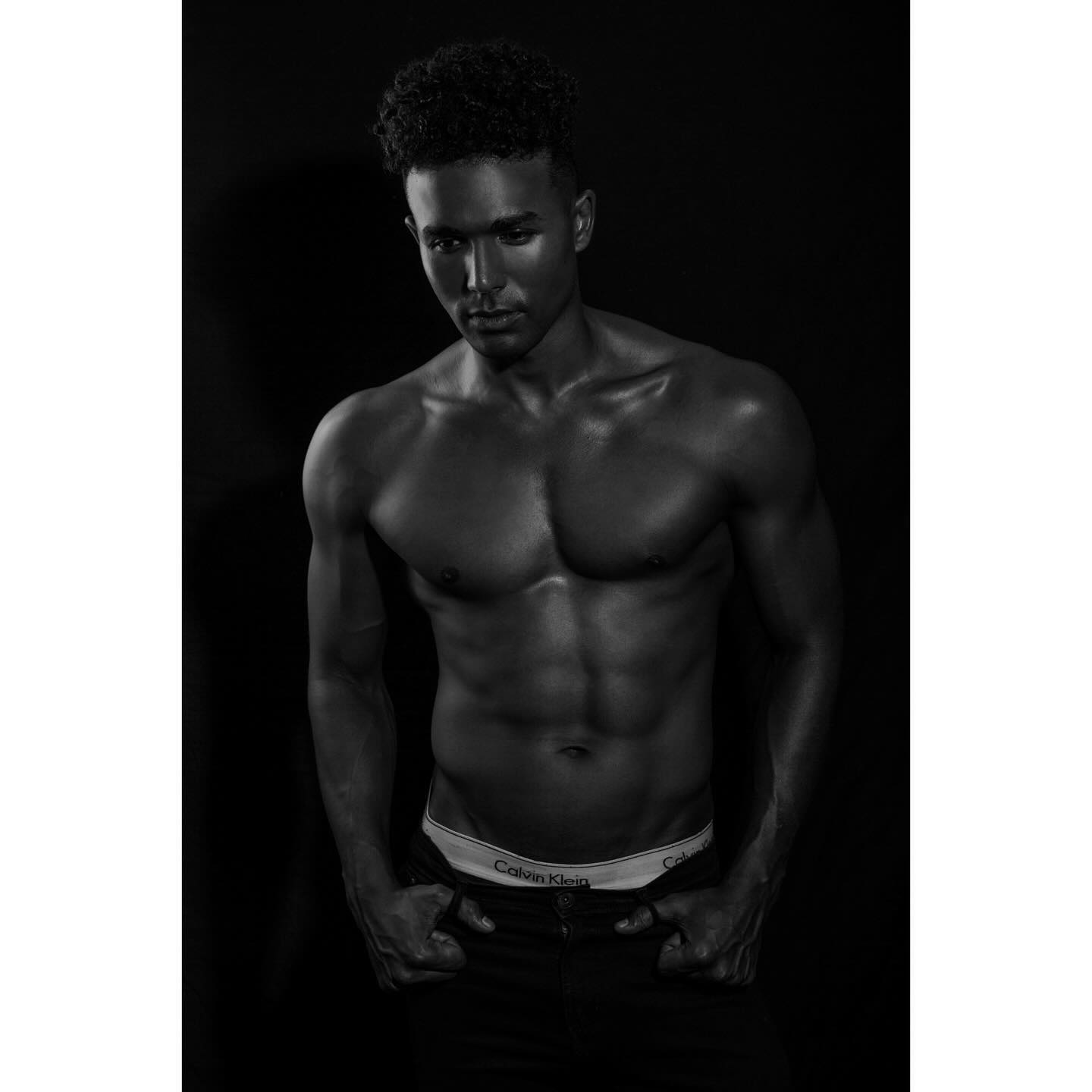 Black and white studio portrait of a shirtless Black man in jeans, posing with hands in pockets, lit by side lighting, with Calvin Klein underwear.
