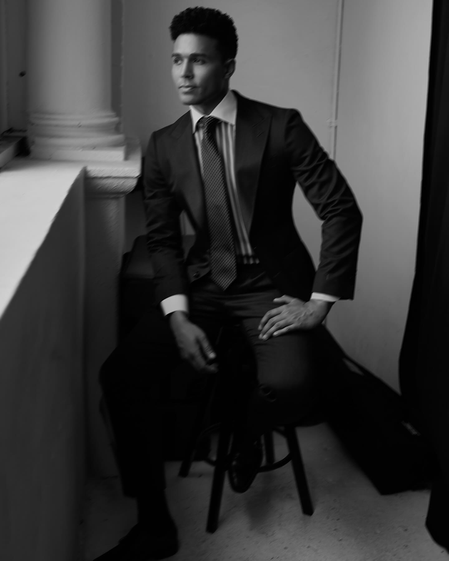 Black and white studio shot of a dark-skinned male model sitting on a stool wearing a suit, tie, and collared shirt looking off camera.