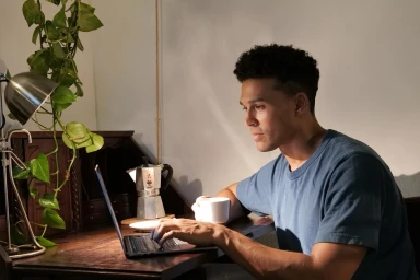 Man wearing a blue t-shirt typing on a laptop at a desk with a potted plant and a coffee cup