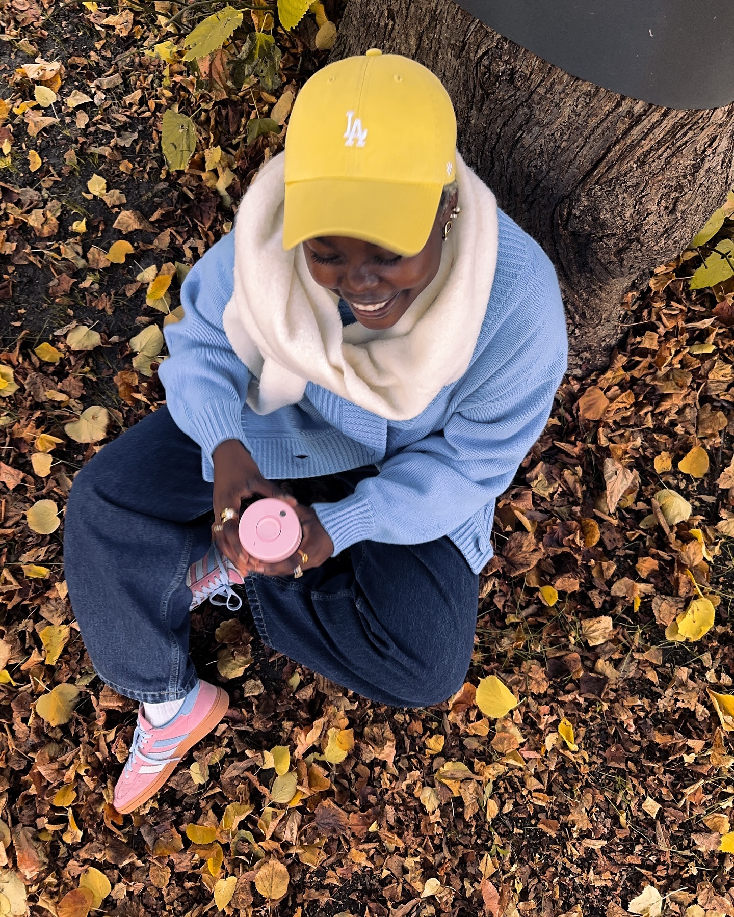 A woman with a yellow cap sits cross-legged on fallen leaves near a tree, holding a pink cup, with a light blue sweater and dark jeans.