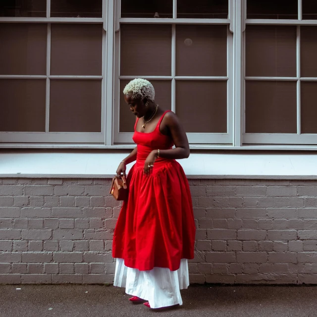 Woman in red dress and white skirt poses near a brick wall with gridded windows, holding a brown handbag and wearing jewelry.