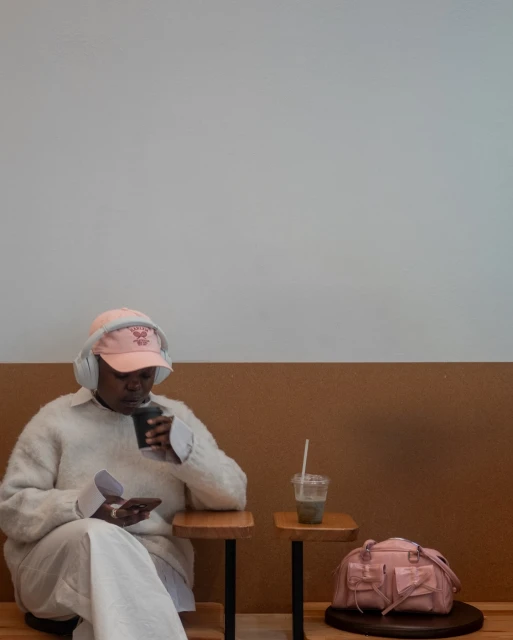 Woman in a pink baseball cap and white headphones sits, sips green juice, and uses her phone in a minimalist interior space.
