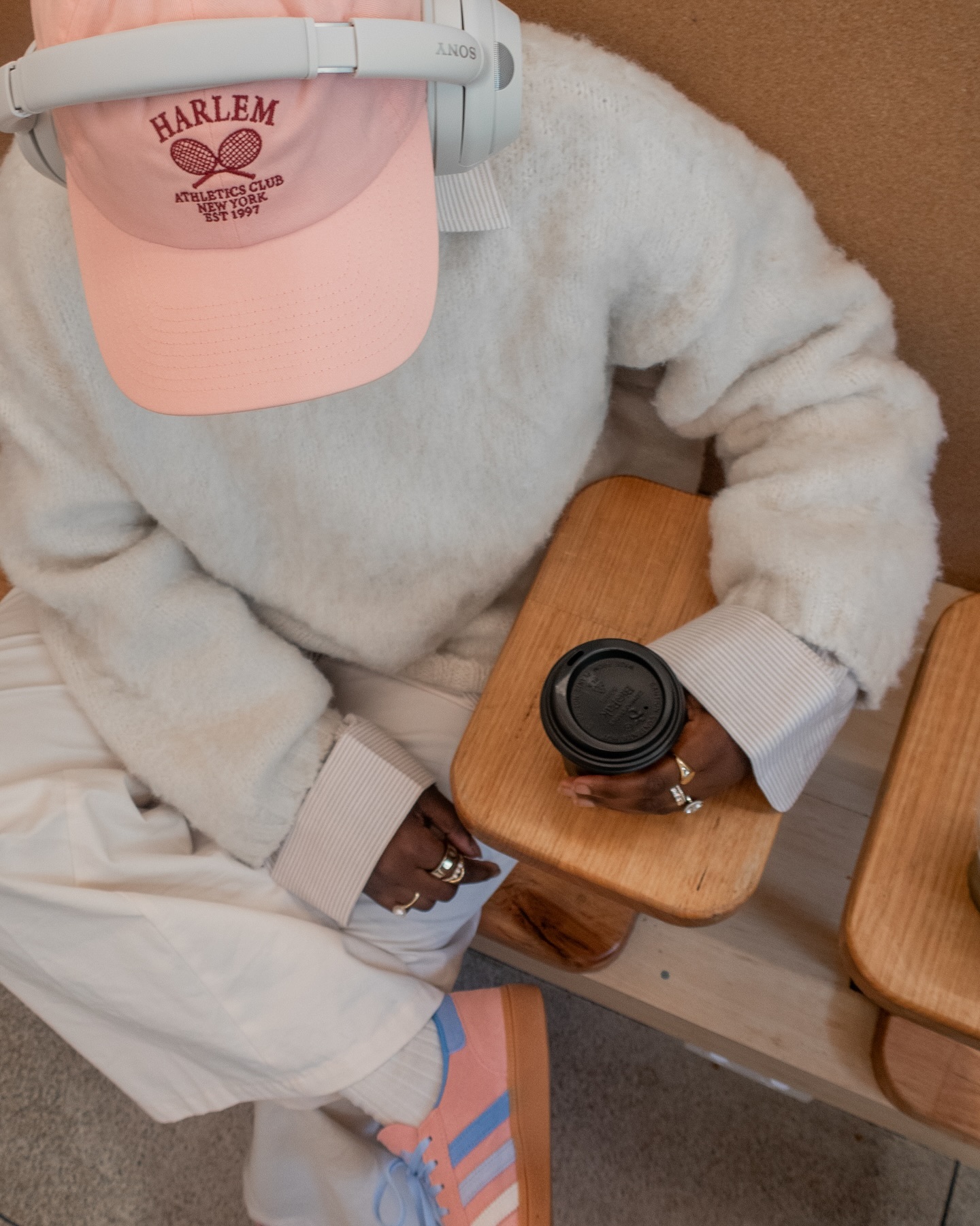 Person sits indoors wearing headphones and a Harlem tennis club hat with a coffee cup, casual sweater, pants, and tennis shoes.