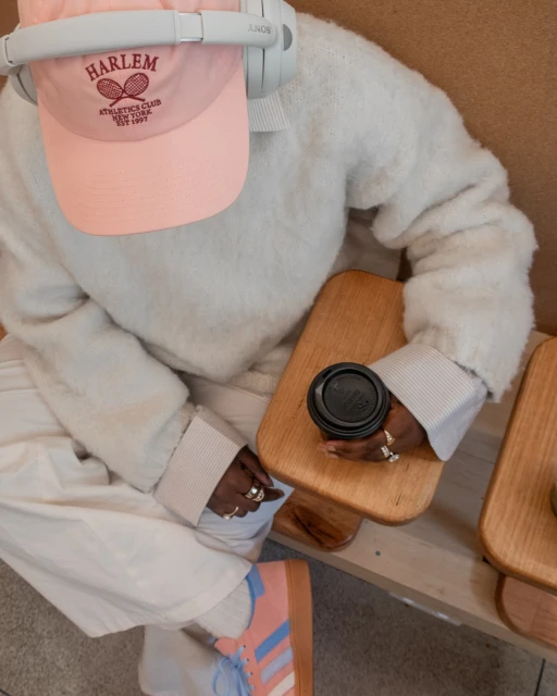 Person sits indoors wearing headphones and a Harlem tennis club hat with a coffee cup, casual sweater, pants, and tennis shoes.