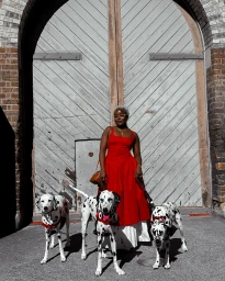 Woman in a red dress standing in front of large wooden doors with three dalmatian dogs on leashes by Tatenda Luna