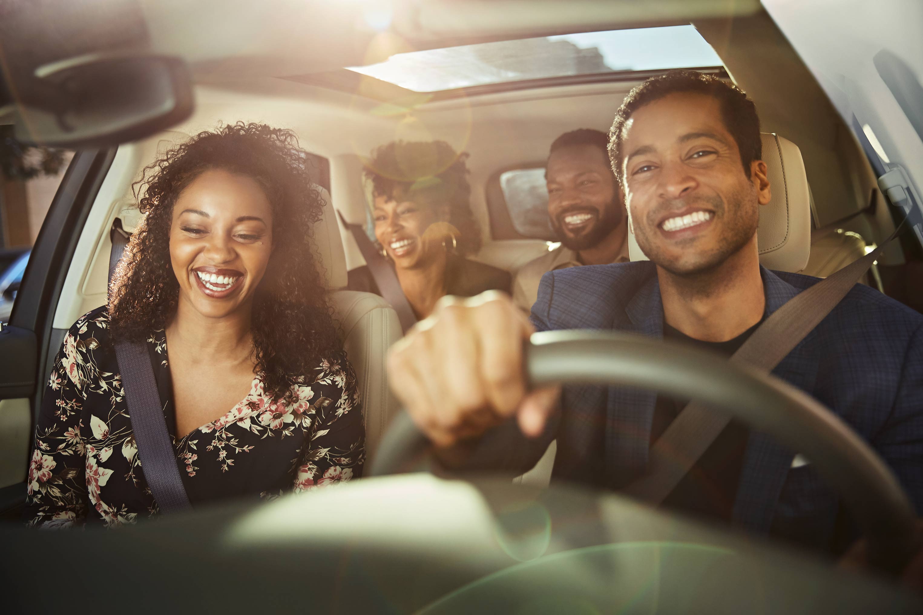 Four adults ride in a car, smiling and laughing in natural light. A man drives while the passengers enjoy the car ride.