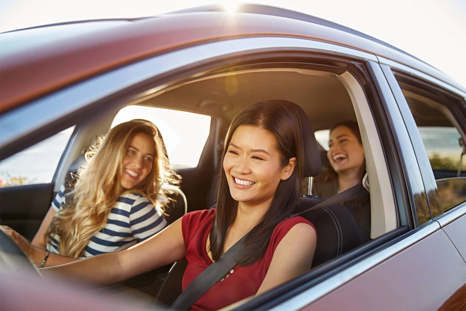 Three smiling women in a red SUV car on a sunny day, one driving, others laughing, enjoying a road trip or vacation.