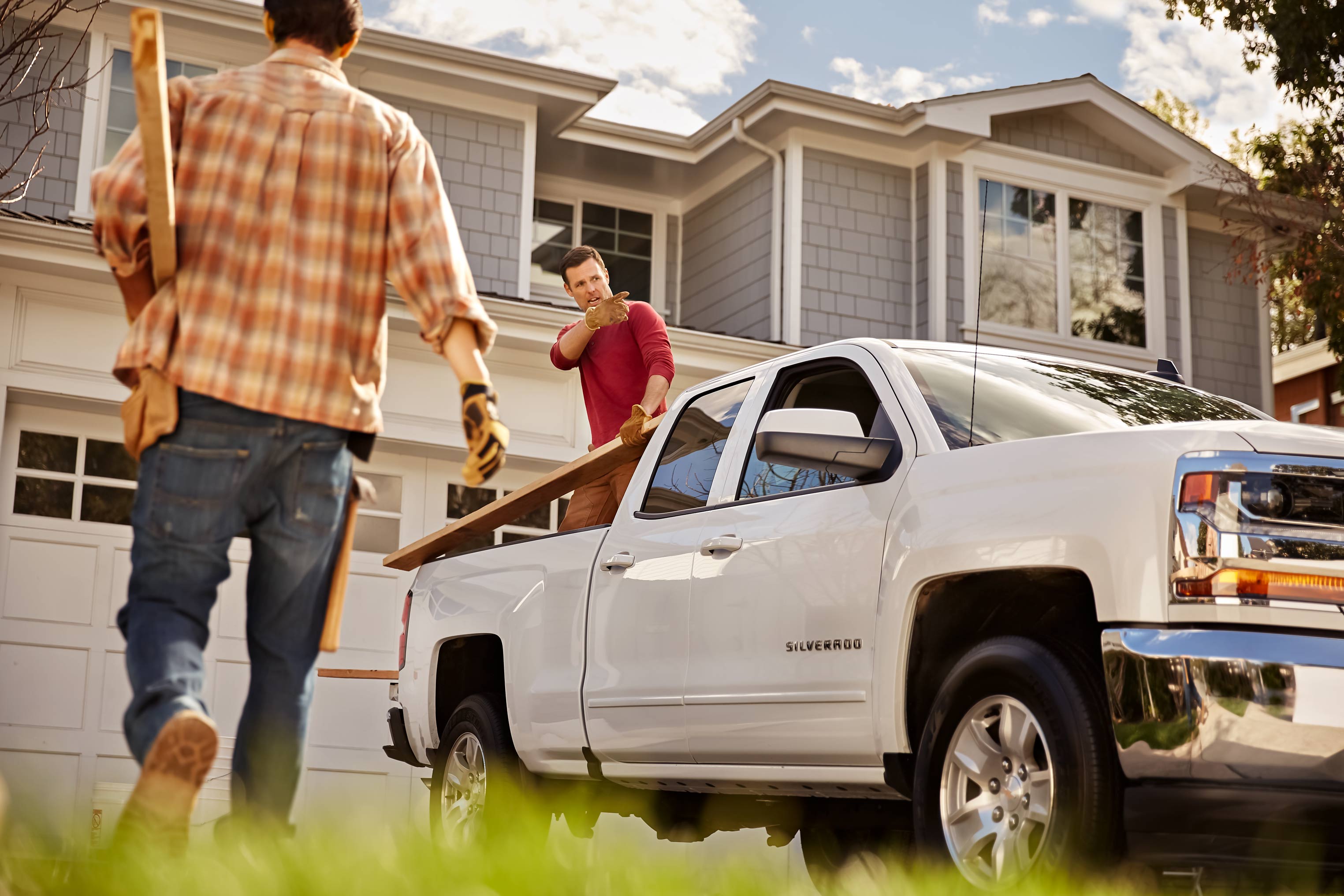Two men loading lumber into a white pickup truck outside a suburban house on a sunny day, showcasing a home improvement scene.