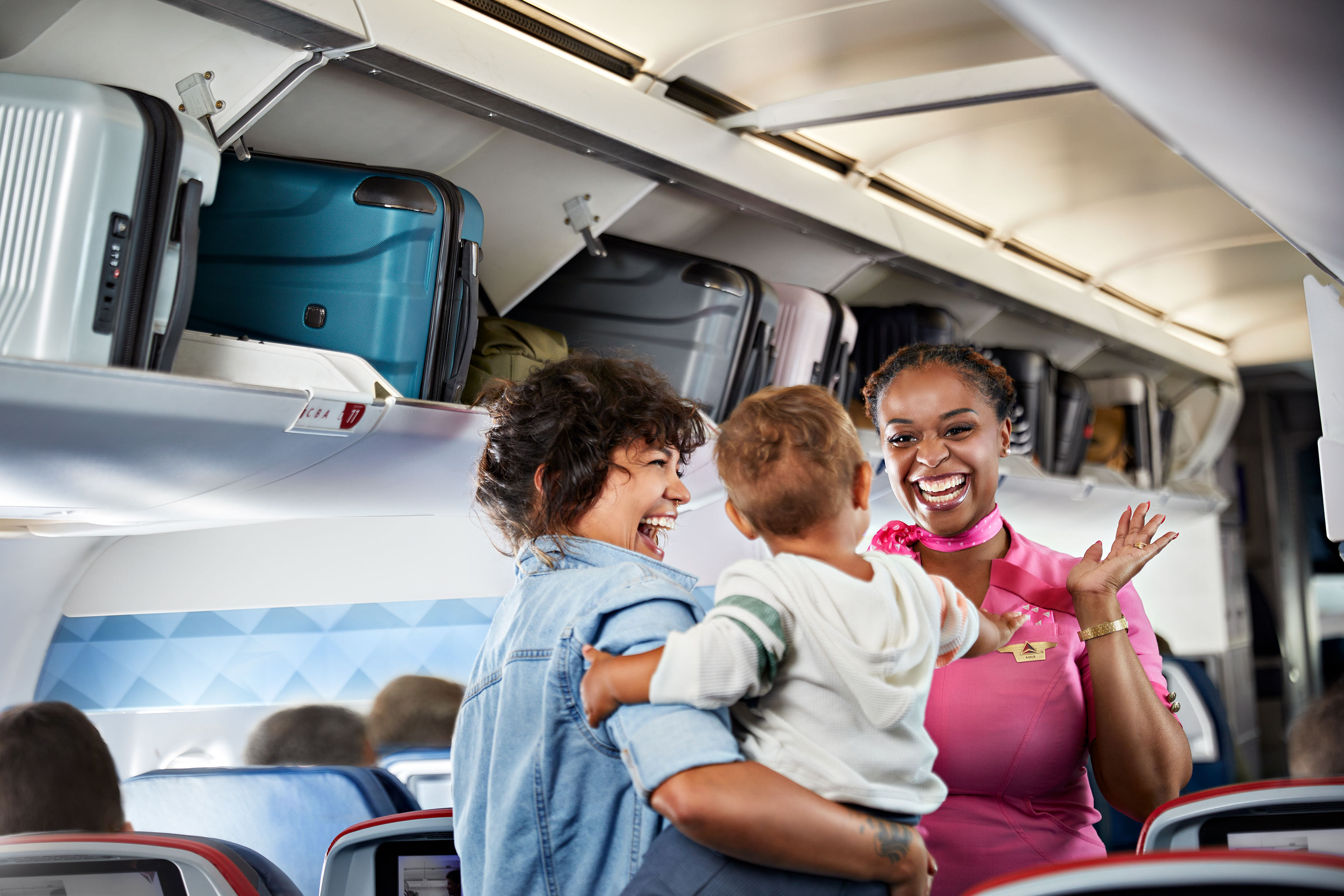On an airplane, a flight attendant greets a mother and child with a smile as they board, with overhead luggage visible.