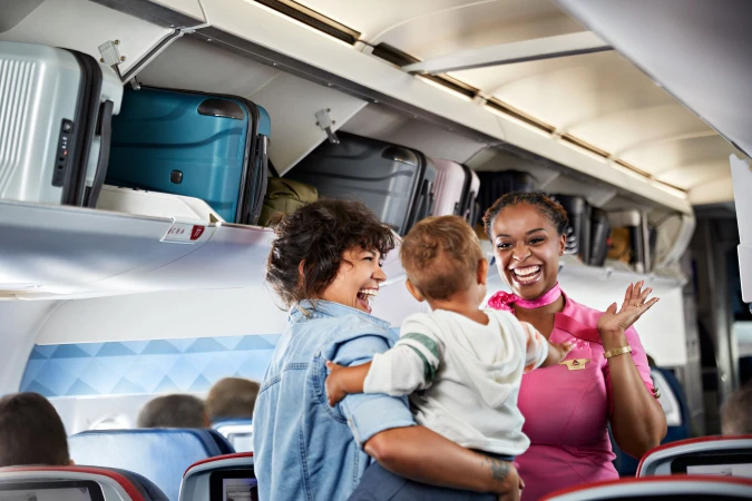 On an airplane, a flight attendant greets a mother and child with a smile as they board, with overhead luggage visible.