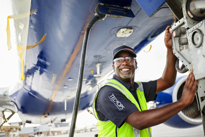 Airplane mechanic performs maintenance, wearing a safety vest and glasses with a blue and white aircraft in the background.