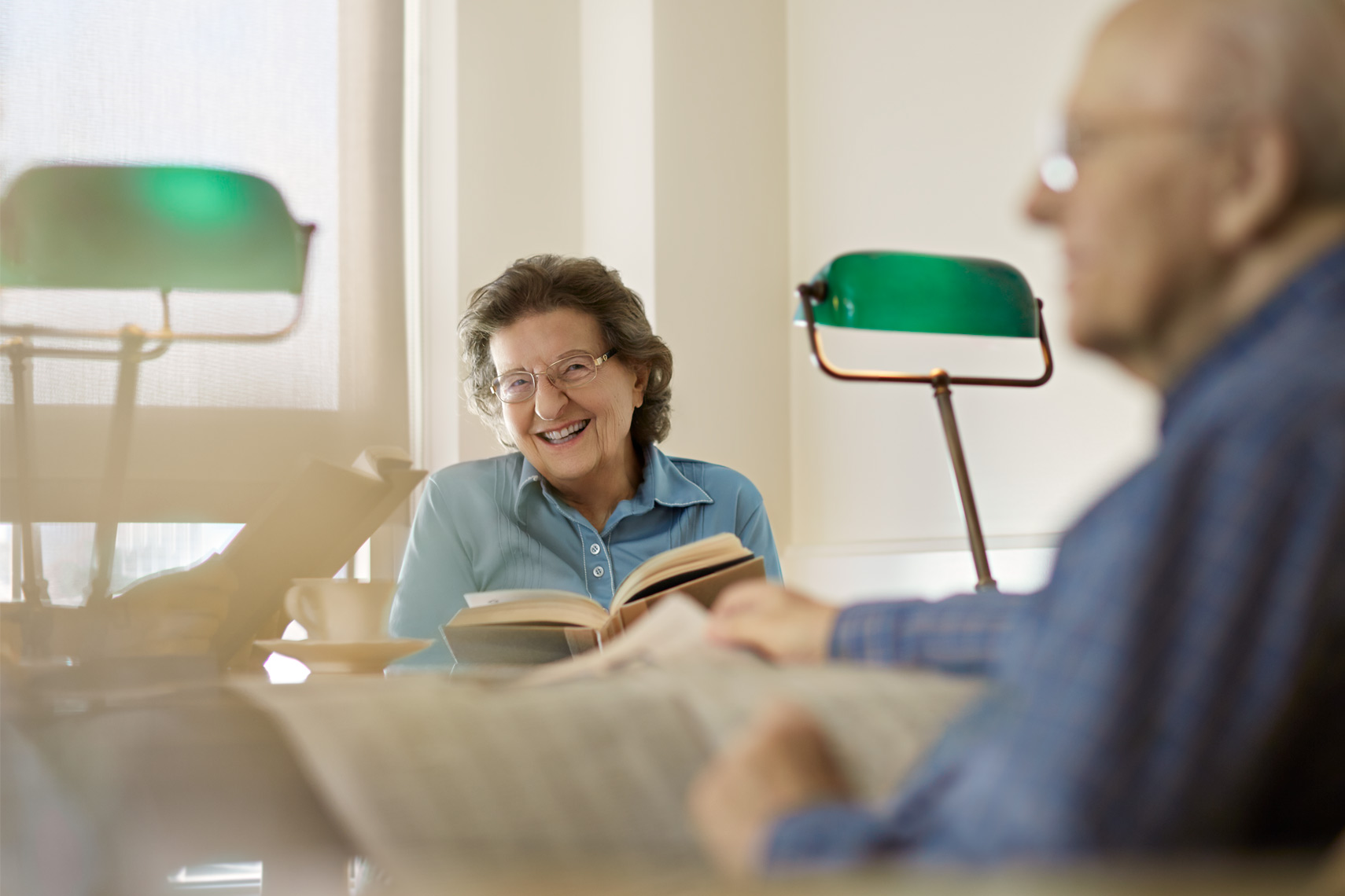 Elderly woman smiles while reading a book next to a lamp. An elderly man reads a newspaper in the background.