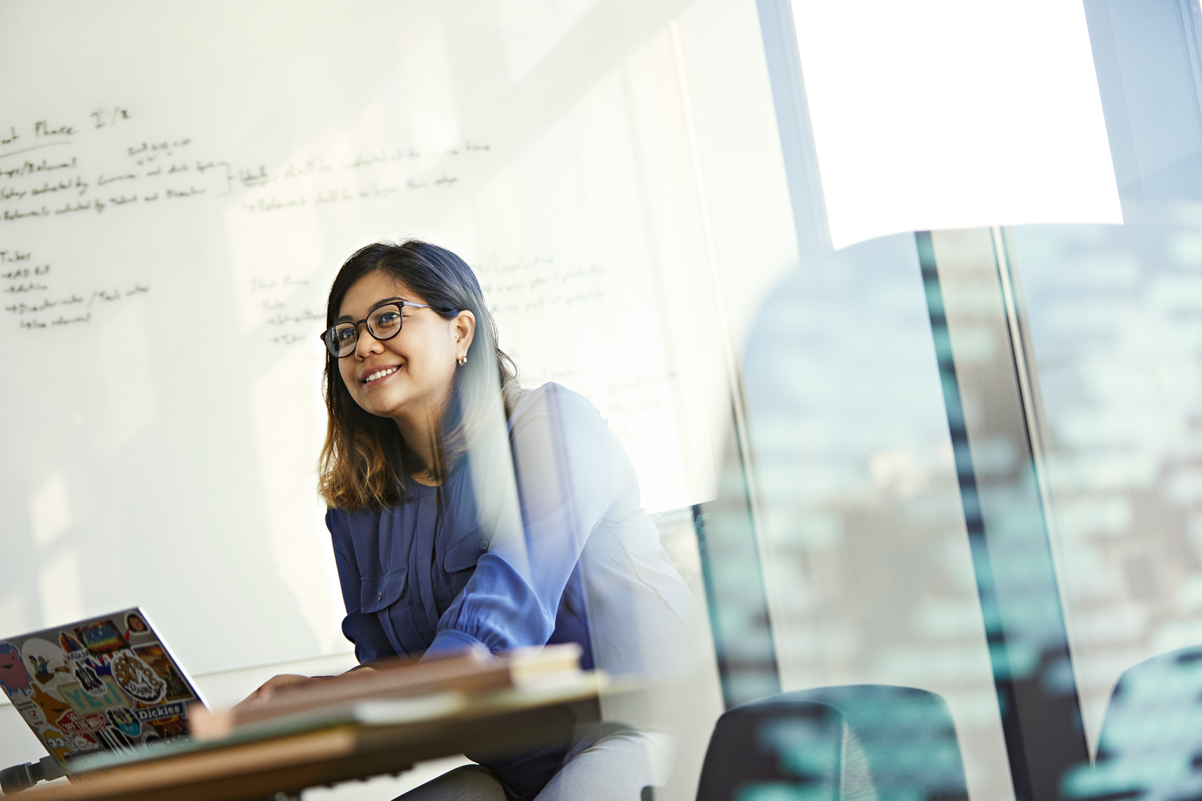 Smiling woman with glasses works on a laptop in an office with a whiteboard. She wears a blue shirt and sits at a table.
