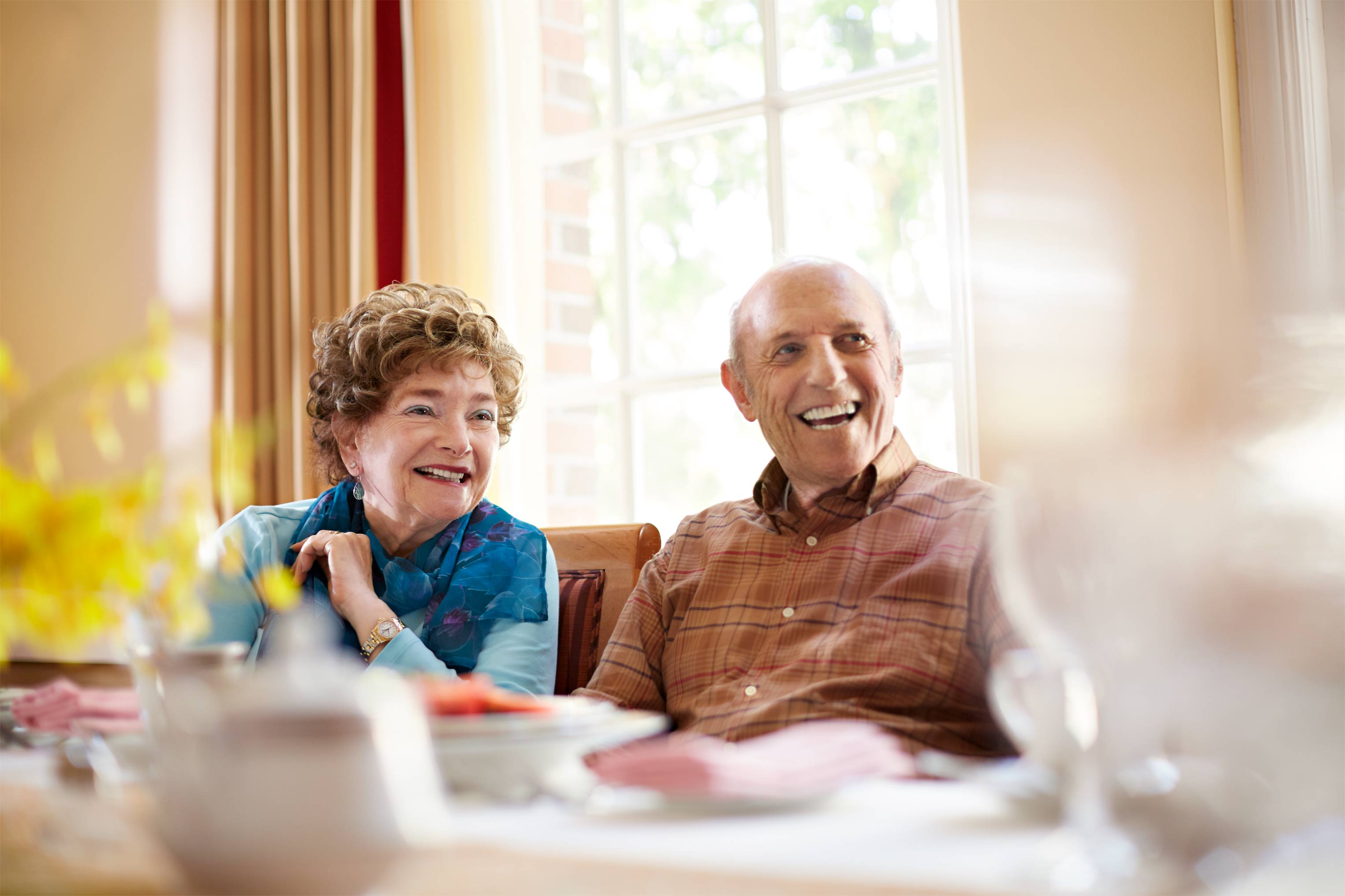Elderly couple smiles while sitting at a table with a bright window in the background. Focus on their faces and joyful expressions.
