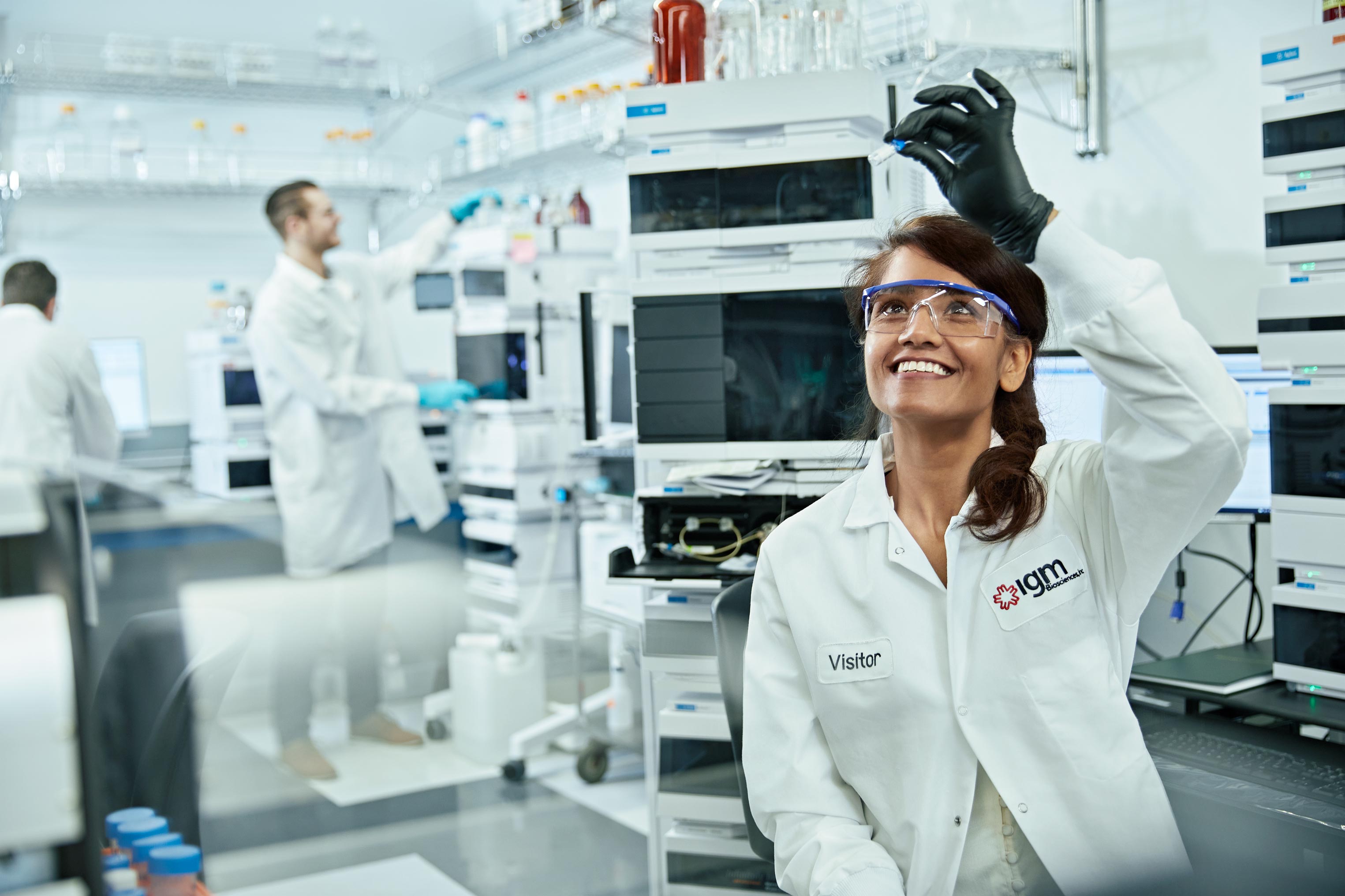 Female scientist in lab coat and safety glasses smiles while holding a vial in a bright, sterile laboratory setting, with lab equipment.