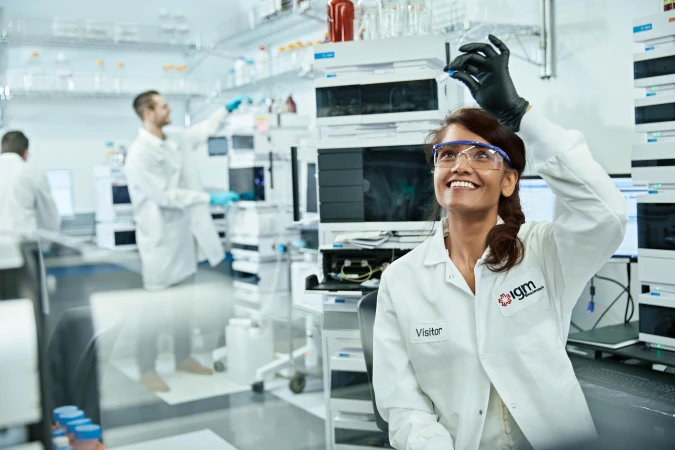 Female scientist in lab coat and safety glasses smiles while holding a vial in a bright, sterile laboratory setting, with lab equipment.