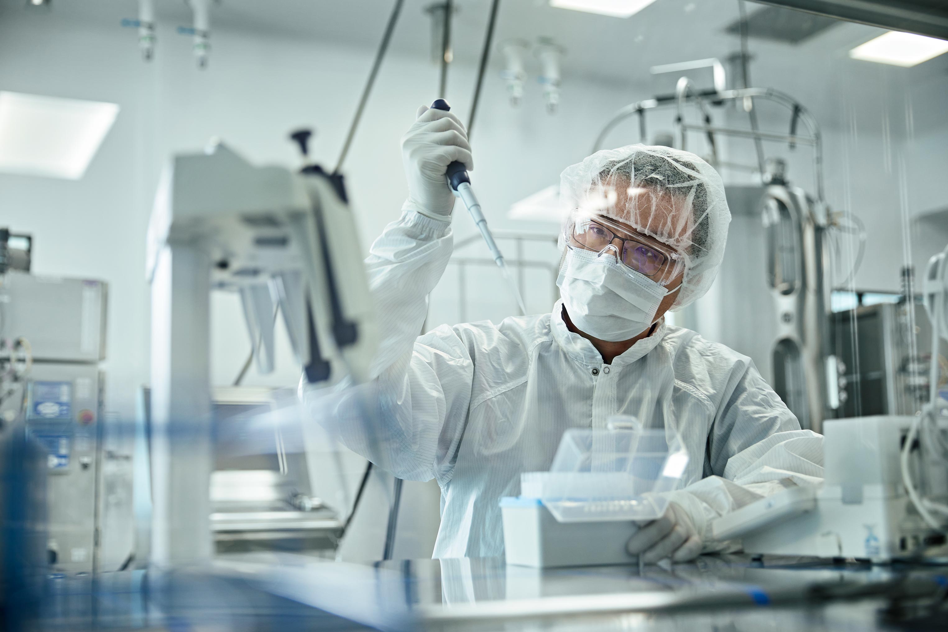 Lab technician in protective suit uses a pipette with laboratory equipment. The bright indoor setting suggests a healthcare environment.