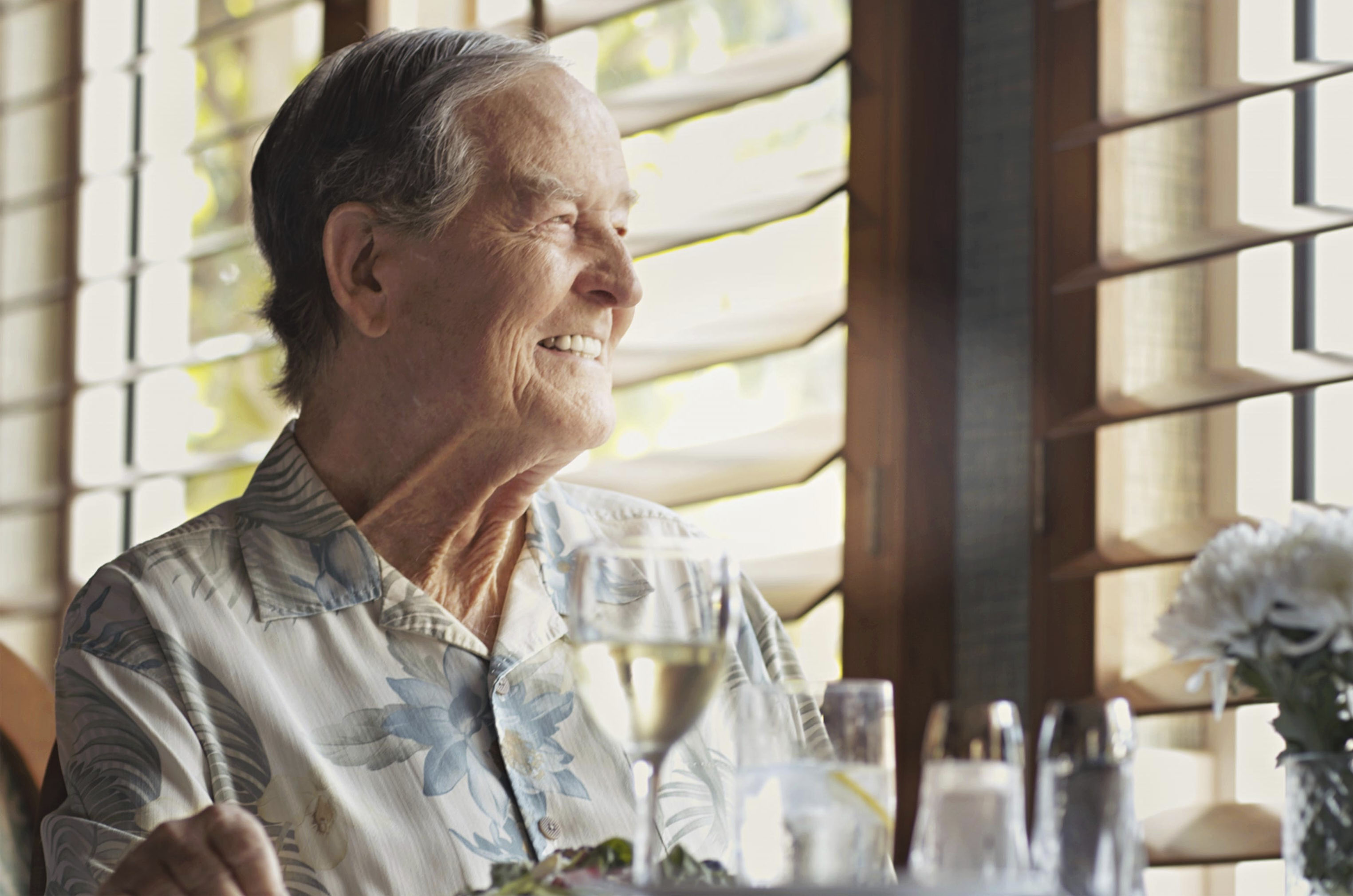 Elderly man smiles while sitting at a restaurant table with a drink, flowers, and window blinds in the background.