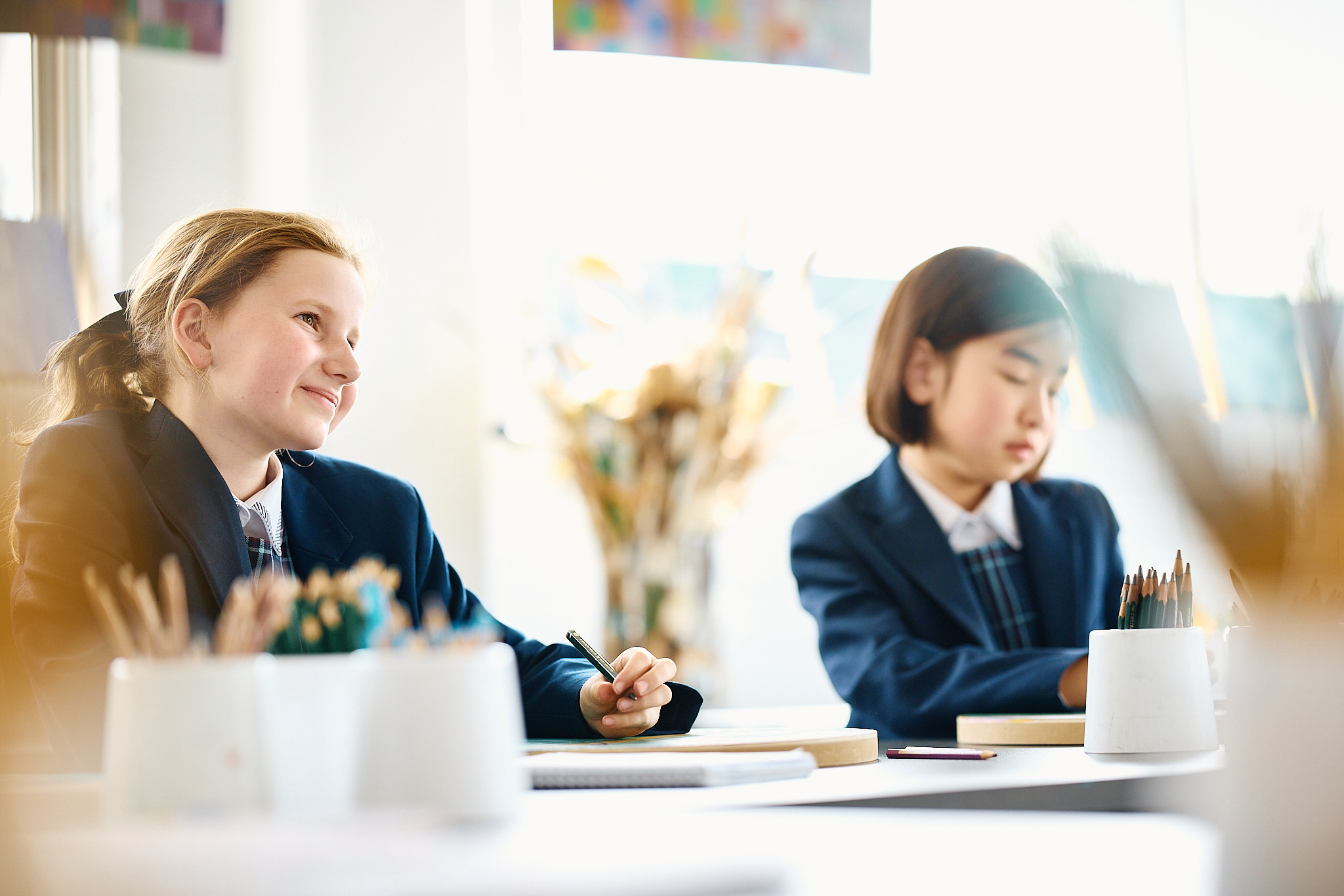 Two schoolgirls in navy blazers sit at a table, one writing and smiling, the other resting with pencils visible in an indoor classroom.