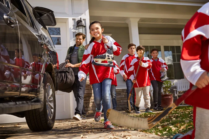 Family arrives at a house, getting out of an SUV. A girl in a hockey jersey carries ice skates, followed by a group of kids with hockey sticks.