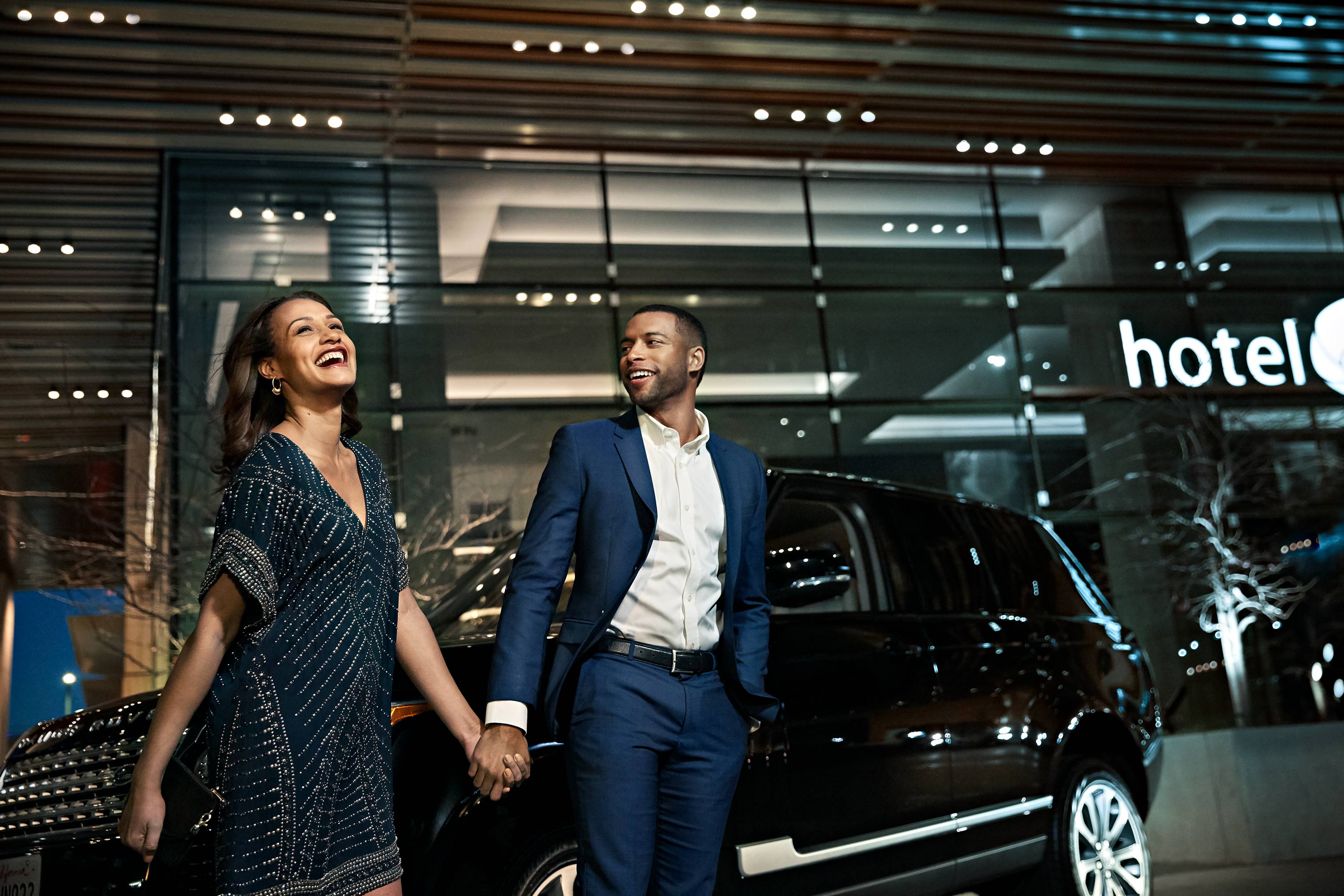 A stylish couple holds hands in front of a luxury SUV and modern hotel at night, smiling and well-dressed, with a contemporary urban setting.
