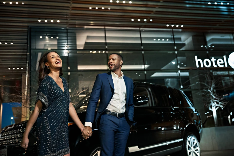 A stylish couple holds hands in front of a luxury SUV and modern hotel at night, smiling and well-dressed, with a contemporary urban setting.