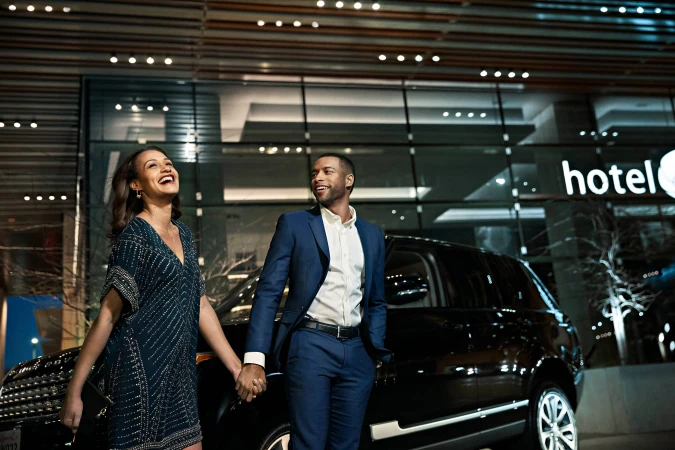 A stylish couple holds hands in front of a luxury SUV and modern hotel at night, smiling and well-dressed, with a contemporary urban setting.