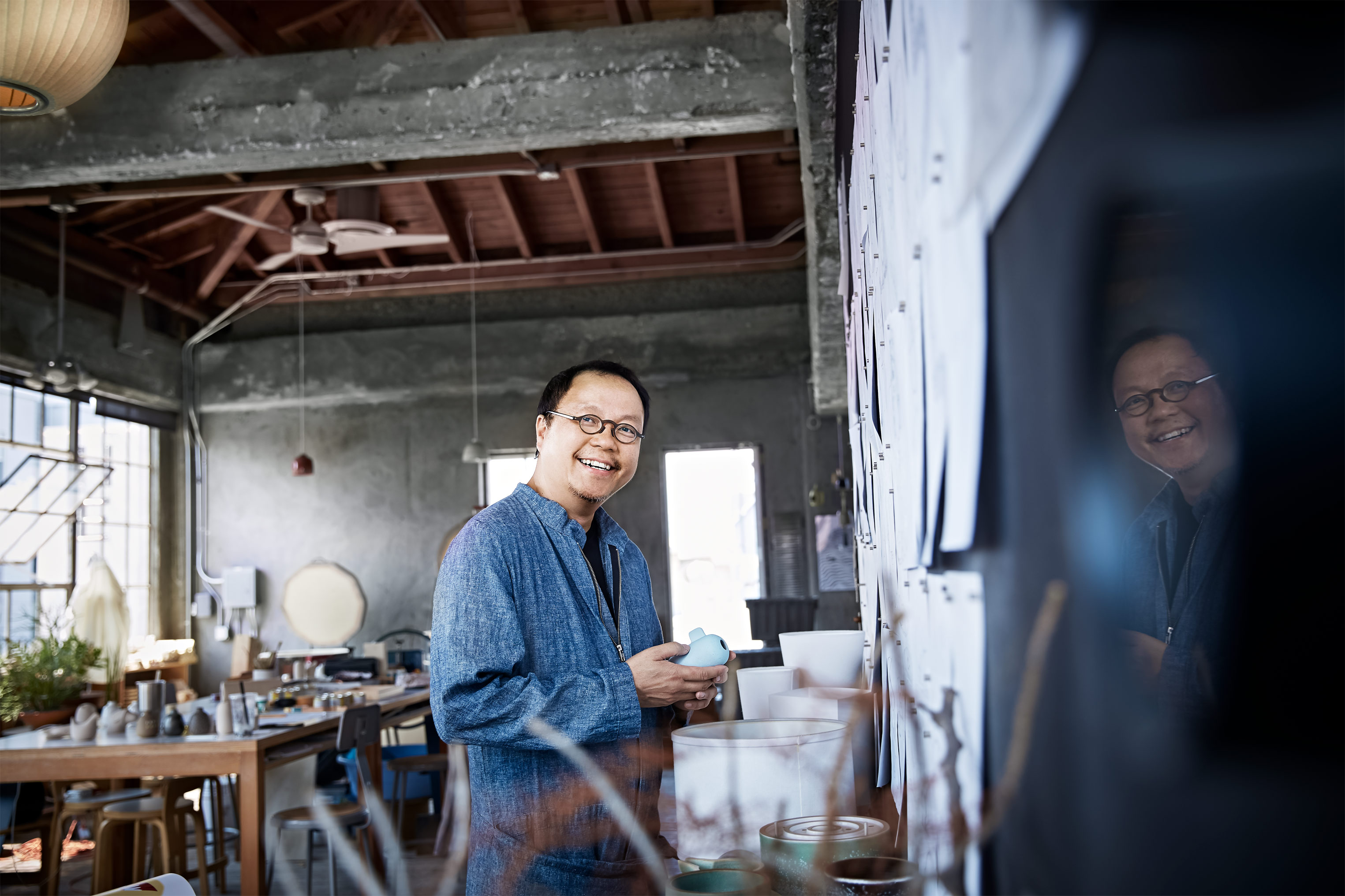 Portrait of a smiling mature Asian man with glasses, wearing a blue shirt and holding a small object, in a sunlit artist's workshop.