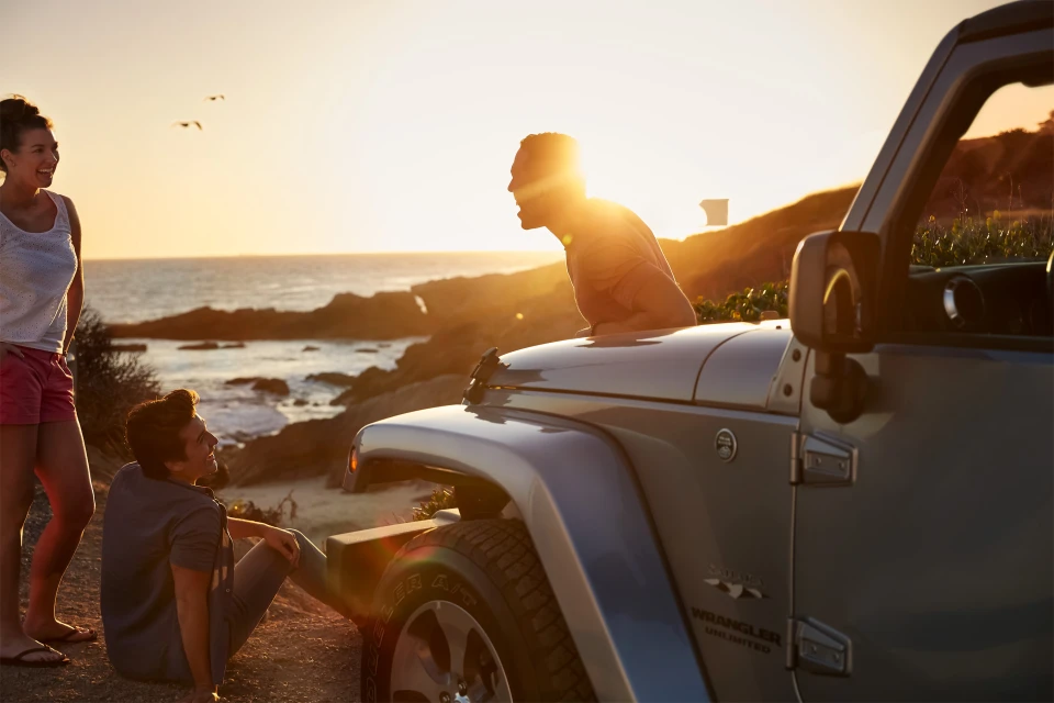 Three people gather around a silver Jeep Wrangler parked on a cliff overlooking the ocean at sunset, creating a scenic and adventurous scene.