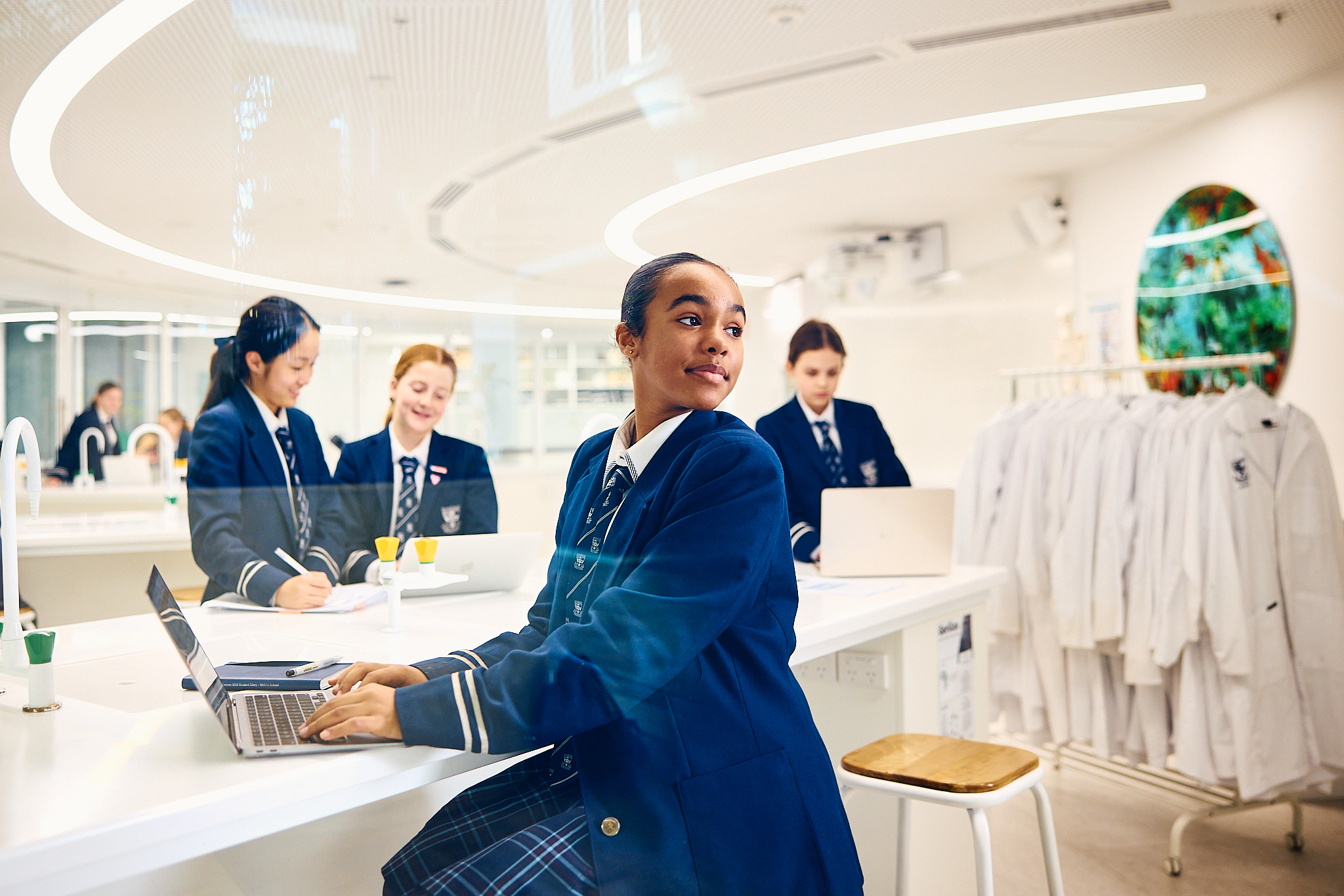 A young woman works on a laptop in a bright, modern science classroom, surrounded by other students in school uniforms.