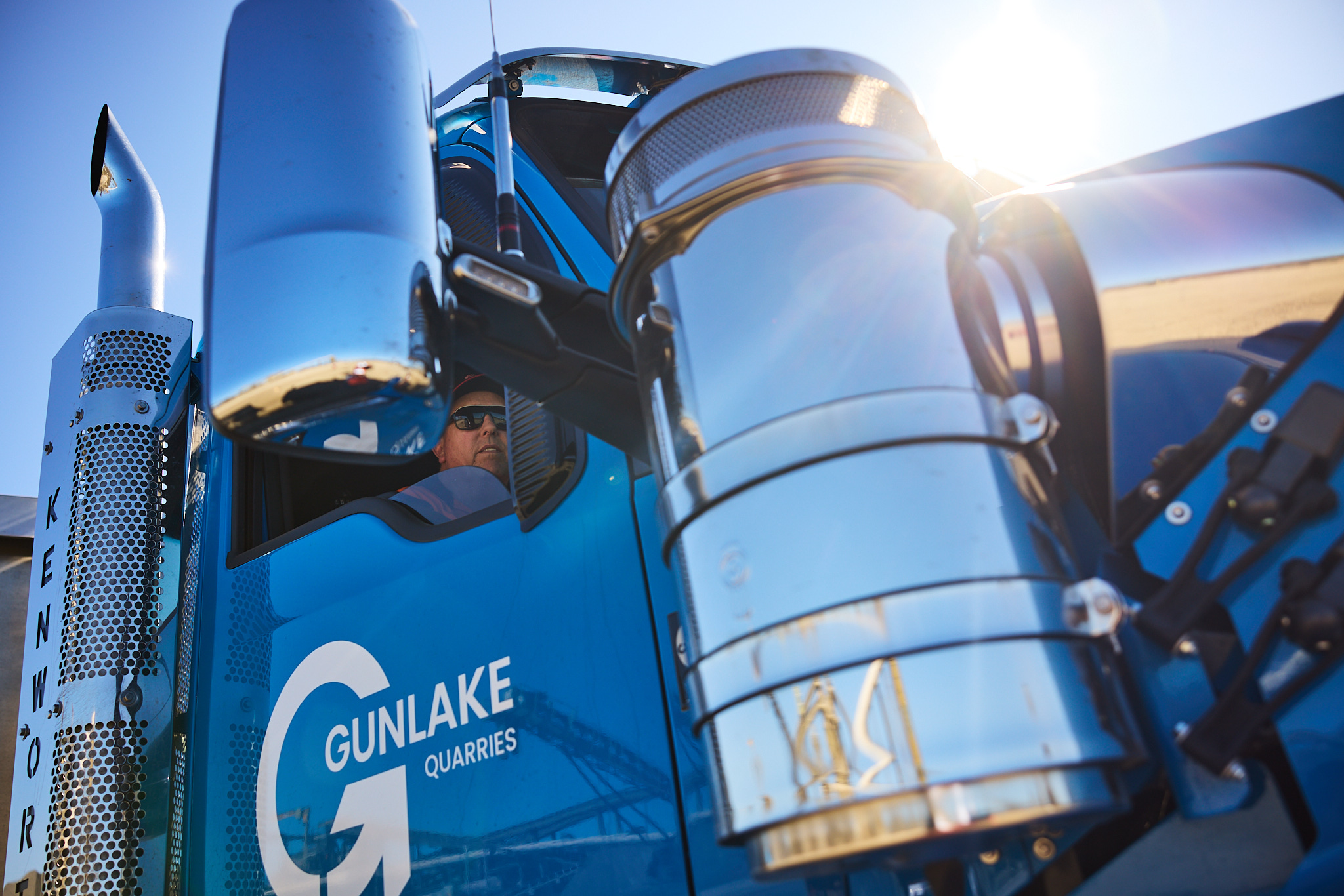 A blue Kenworth semi-truck with a driver wearing sunglasses visible through the window, and the company logo "Gunlake Quarries" on the door.