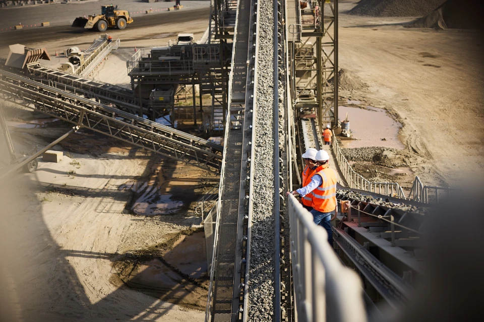 Two construction workers wearing hard hats and orange vests are standing near conveyor belts at an industrial site on a sunny day.