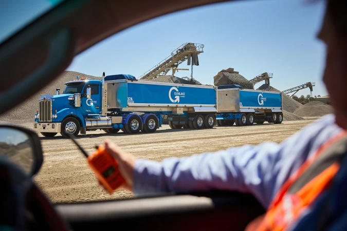 Blue Gunlake Group semi-truck hauling gravel along a dirt road, observed from inside a vehicle on a sunny day with blue skies.