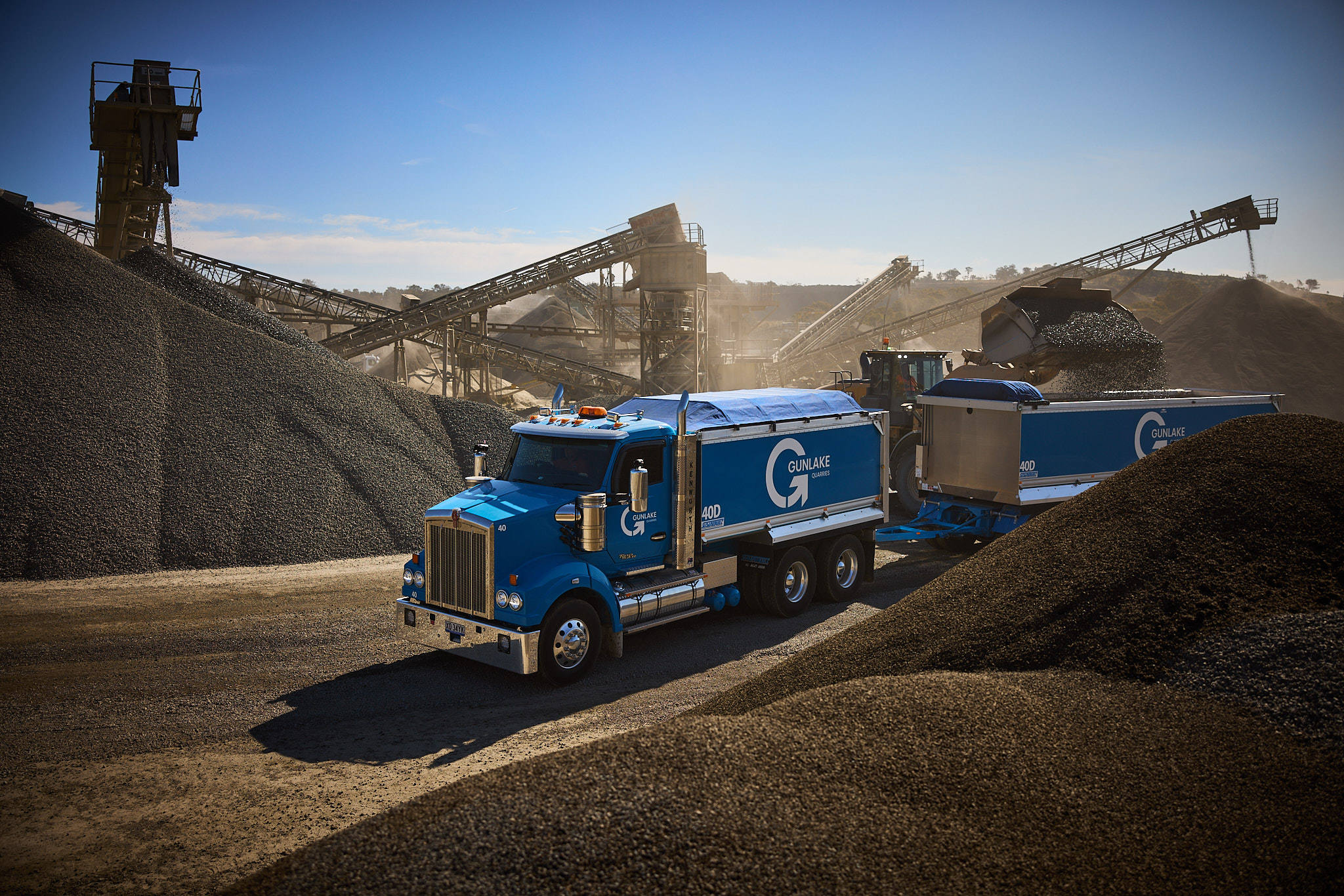 Blue Gunlake Group dump truck parked on a dirt road at an outdoor industrial rock quarry on a sunny day with rock piles visible.